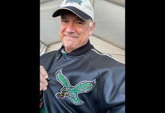 A selfie of Macnow. He is an older man with white hair. He wears a white sports cap and a black letterman jacket. He pulls at his jacket lapel to show an image of a green eagle with outstretched wings holding a football in its talons. 