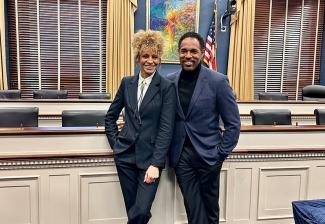 Hurd and George stand in an office. Both are dressed in dark suits. Behind them is a long table with office chairs and windows with gold drapes; a painting and an American flag. A blue, star-patterned carpet is underfoot. 