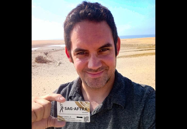 An individual holds their SAG-AFTRA membership in their hand. They are standing outside in a desert-like area. They wear a charcoal-colored shirt. The card is white with the SAG-AFTRA logo in black and gold curving brushstrokes in the upper right- and lower left-hand corners.