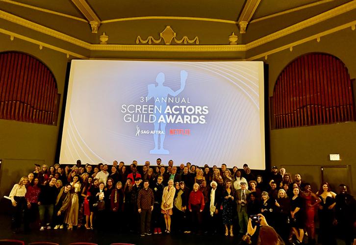 A large group takes a photo on stage in a movie theater. Behind them on a projector screen is the title card for the 31st SAG Awards ceremony.