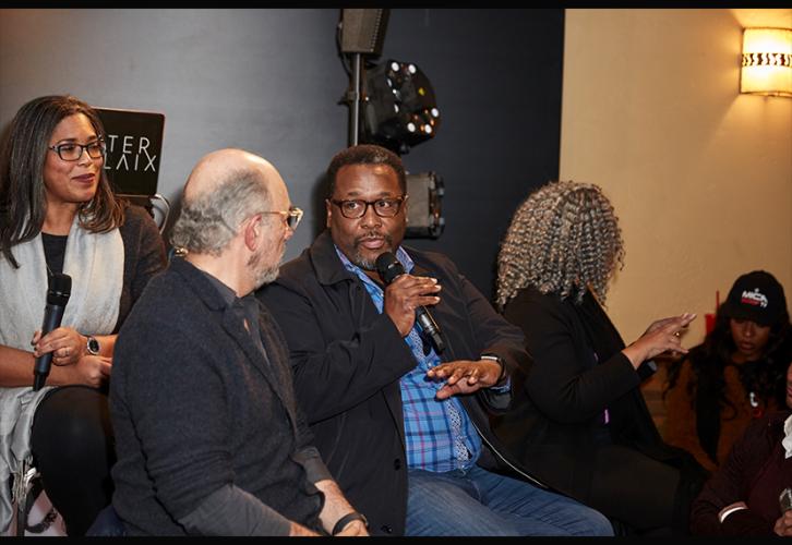 A seated African-American male, center, turns and speaks to a Caucasian male.