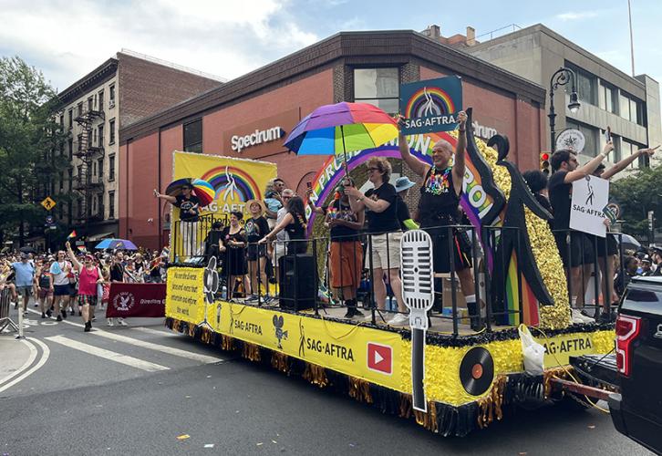 A group of people ride a yellow parade float as the float turns a street corner in New York City. They hold rainbow-paneled umbrellas and SAG-AFTRA signs with a rainbow on a teal background.