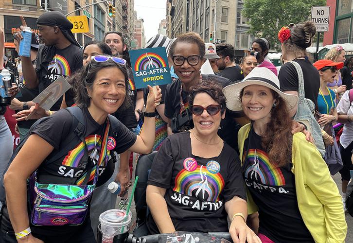 A group of four people pose together facing the camera. They are all wearing black T-shirts with a rainbow and a white SAG-AFTRA logo on the front. 