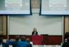 A wide shot of Fowler in a lecture hall. Fowler is located in the background behind a podium. Above her are two projector screens that showcase a slide from her presentation. The audience is seated in the foreground, facing away from the camera.